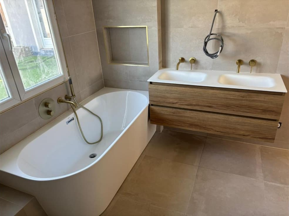 Salle de bain en grands carreaux beige aspect pierre avec baignoire adossée, niche à profilé doré et meuble double vasque, Meyzieu