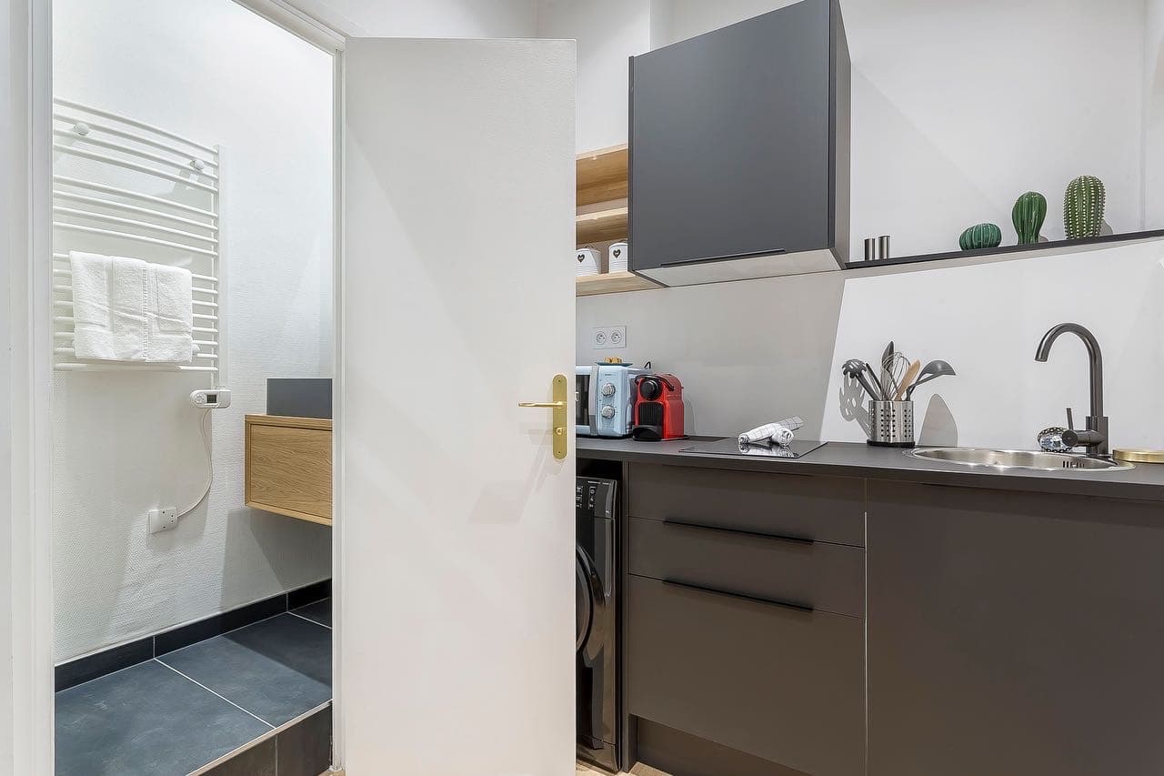 Kitchenette grise ouverte sur une petite salle d’eau au sol carrelé gris anthracite à joints fins, studio à Bourgoin-Jallieu