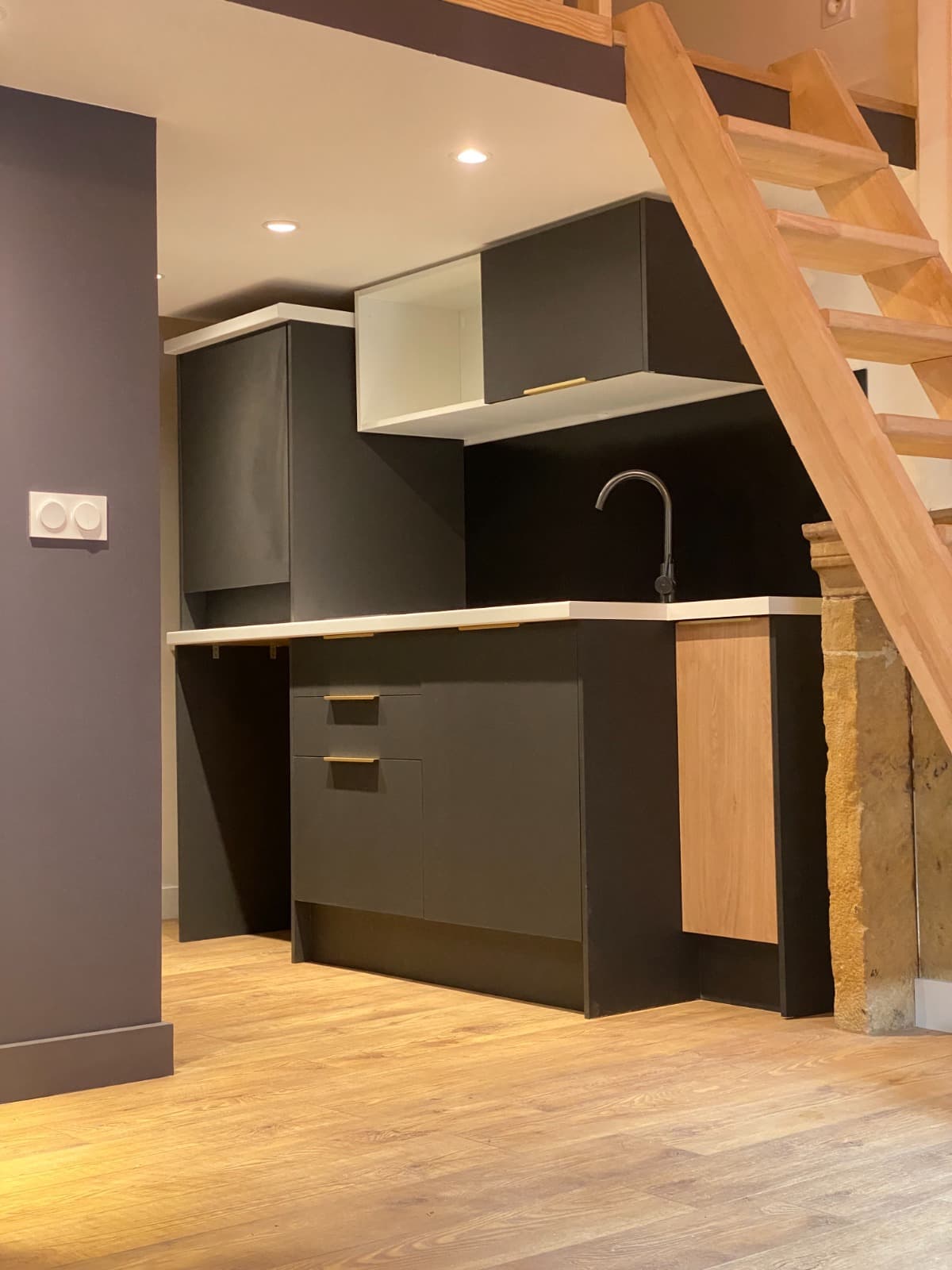 Kitchenette noire mate à poignées laiton et plan blanc sous la mezzanine, près d’une cheminée en pierre, dans un studio à Lyon