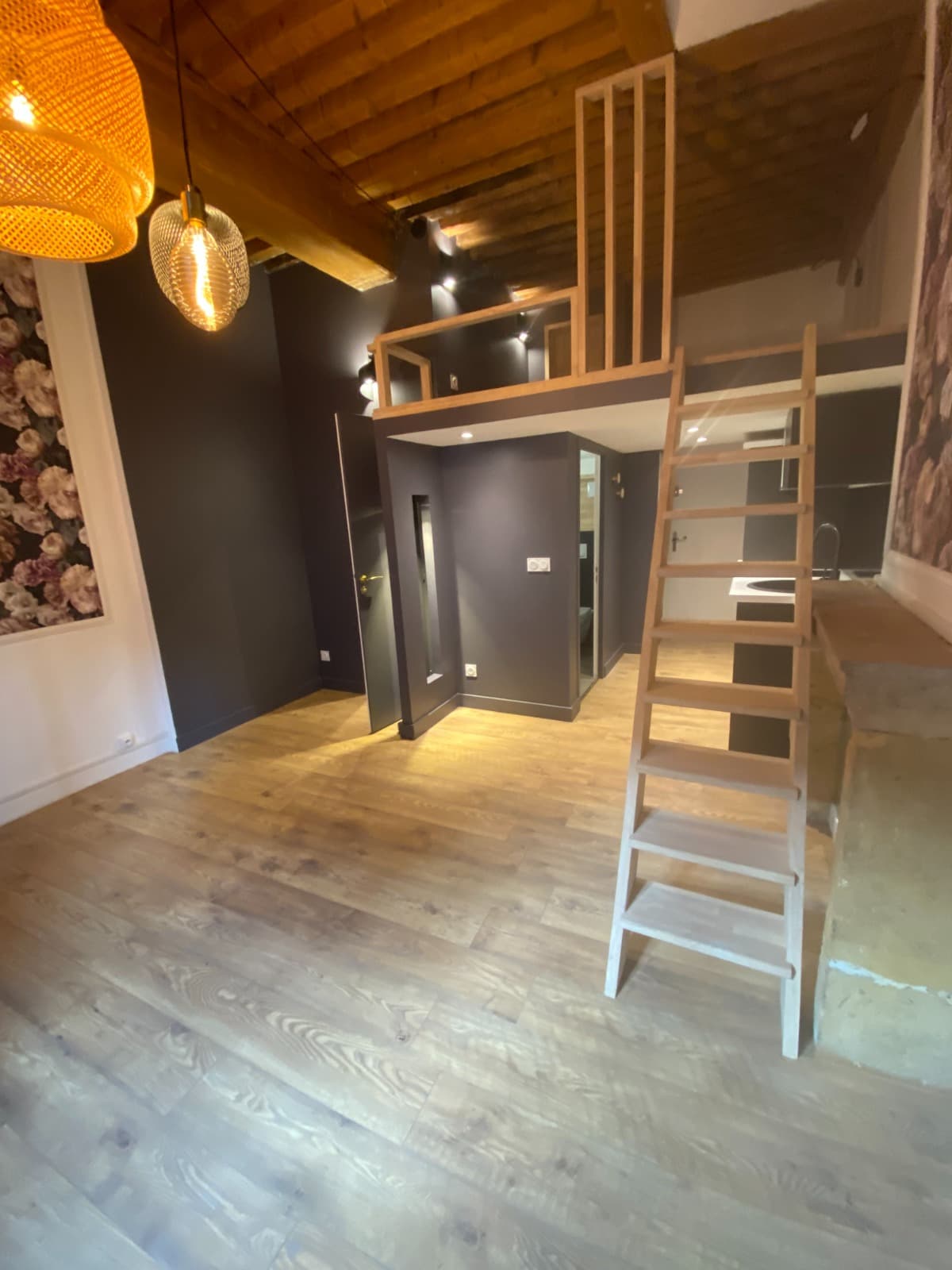 Studio rénové à Lyon : mezzanine en bois clair sous poutres anciennes, échelle de meunier et volume en cloisons peintes anthracite