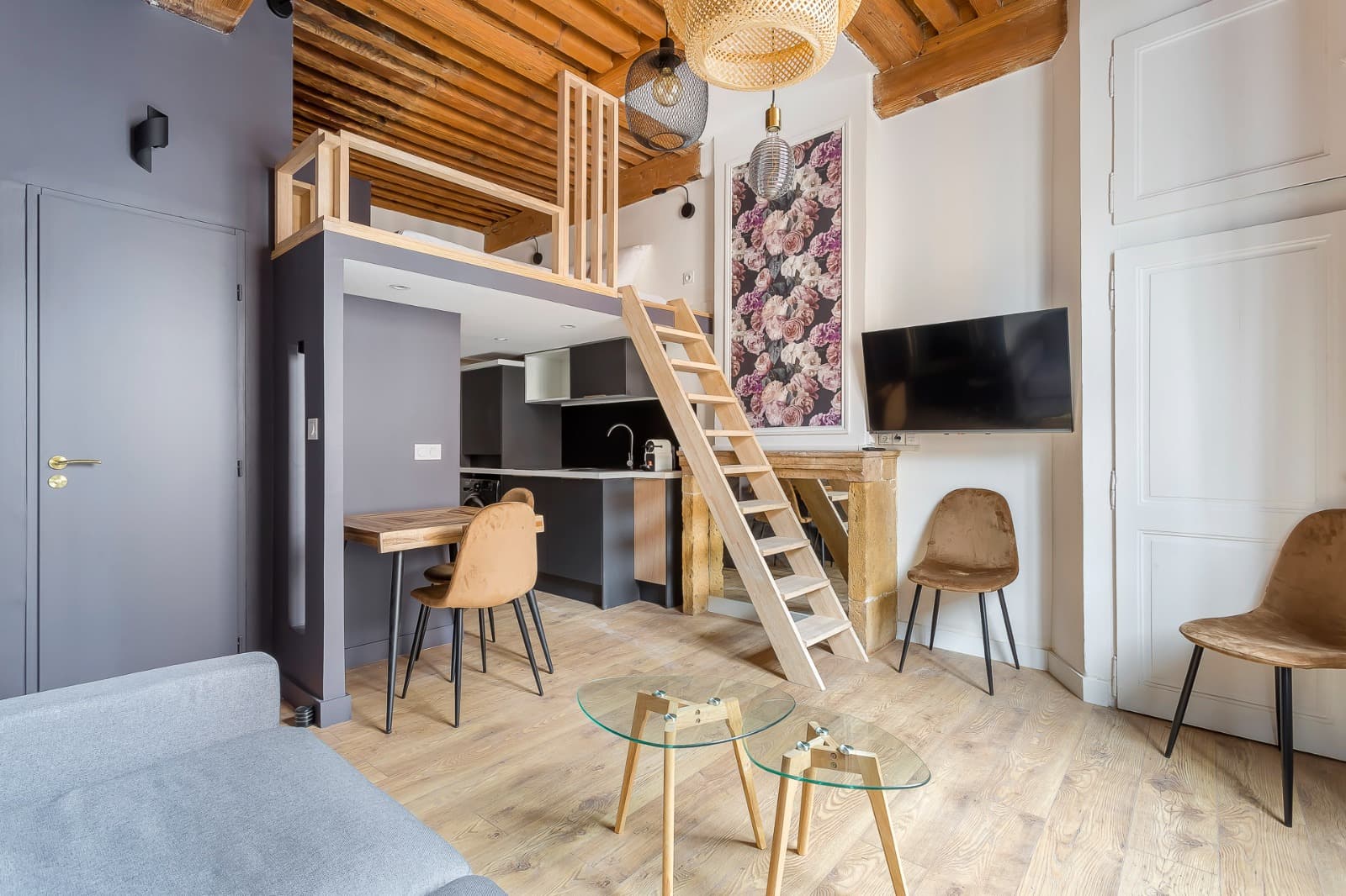 Studio meublé à Lyon : mezzanine à garde-corps bois au-dessus d’une cuisine anthracite, solives apparentes et papier peint floral